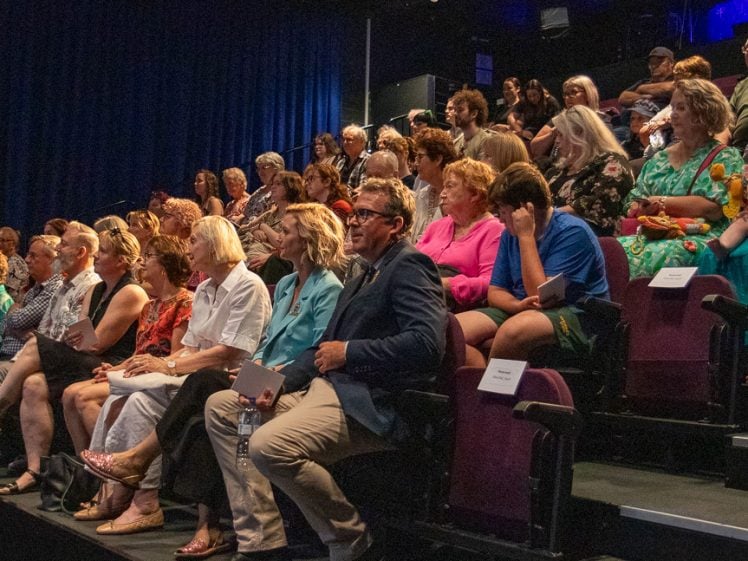 Image of A diverse audience of adults and children sits in tiered seating inside a dimly lit theatre, attentively watching an event on stage. Some people hold programmes, and a child in the front row holds a doll.