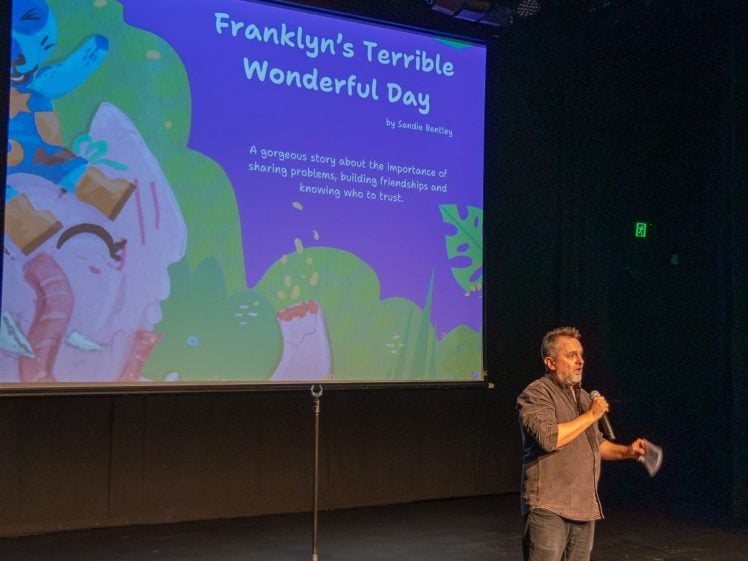 Image of A man stands on stage holding a microphone and paper, speaking in front of a large screen displaying a colourful book cover titled “Franklyn’s Terrible Wonderful Day” by Saskia Bexleys, featuring cartoon animals.