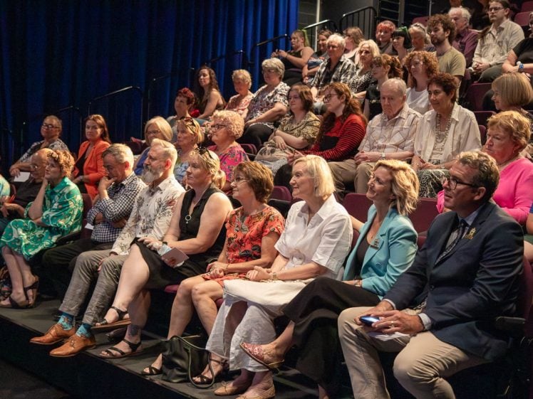 Image of A diverse audience of adults sits attentively in tiered seating, watching an event or performance on stage. The background features dark blue curtains and the atmosphere appears engaged and focussed.