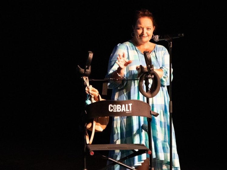Image of A woman in a blue and white dress stands on stage behind a microphone, smiling and gesturing with her hand. In front of her is a black mobility walker labelled “COBALT,” with a bag hanging from it.