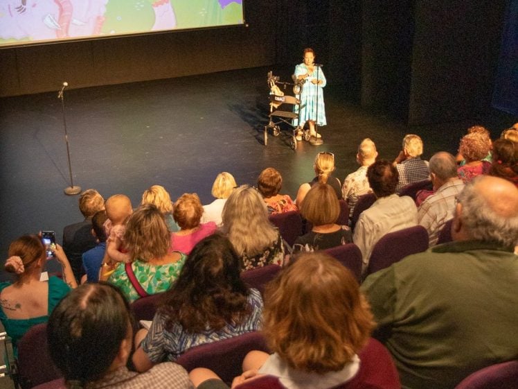 Image of A woman stands on stage with a microphone and props, presenting to a seated audience in a theatre. A colourful screen behind her displays the title Franklyn’s Terrible Wonderful Day.