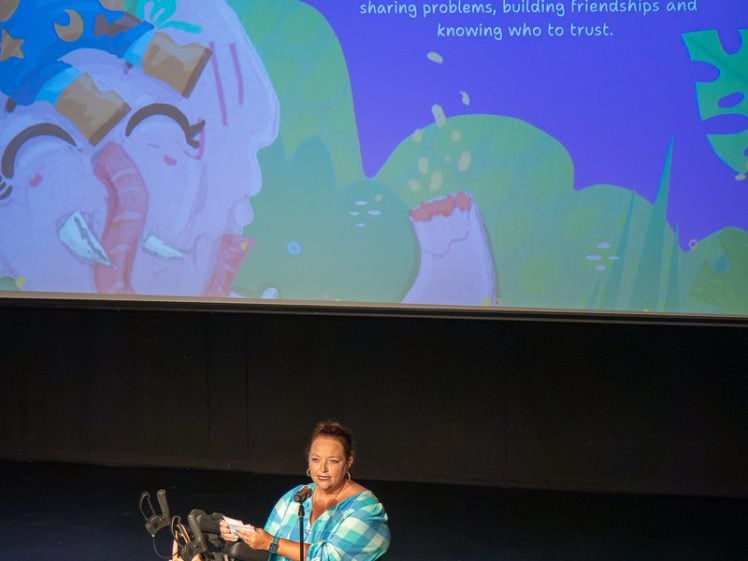 Image of A woman in a blue checked dress stands on stage with a zimmer frame, speaking to an audience. Behind her is a colourful projection titled Franklyns Terrible Wonderful Day by Sandie Bentley, with an illustration of animals.