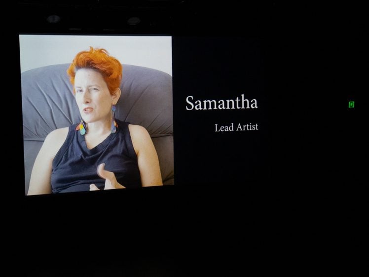 Image of A video screen shows a person with short red hair sitting in a chair, gesturing with one hand. White text beside them reads, Samantha, Lead Artist, on a black background.