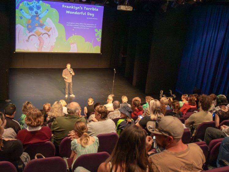 Image of A man stands on stage speaking to an audience in a theatre. A colourful screen behind him reads Franklyns Terrible Wonderful Day, featuring cartoon characters and event details. Audience members watch attentively from purple seats.