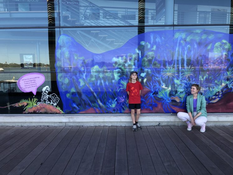 Image of Two children pose in front of a vibrant, underwater-themed mural on a glass wall. One stands in a red shirt, while the other crouches in a teal jacket. The mural features blue, purple, and green aquatic plants.