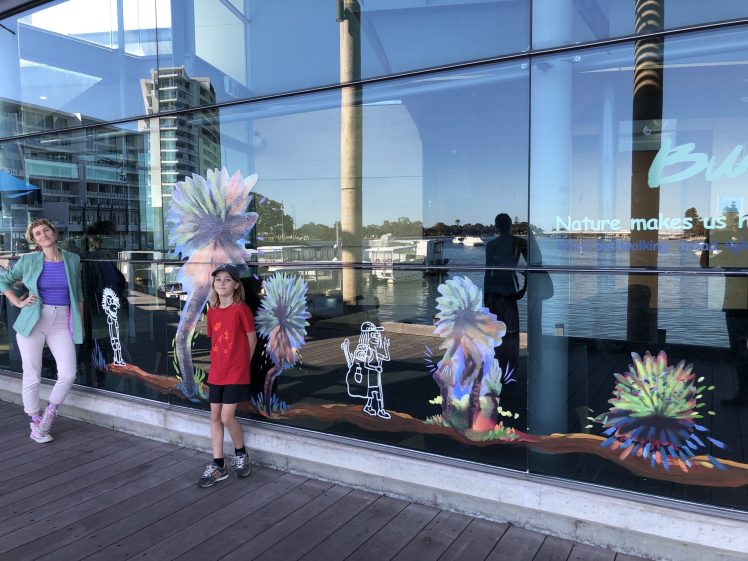 Image of A woman and a child stand smiling on a wooden walkway in front of a large glass window decorated with colourful abstract art and white line drawings. Reflections of water and buildings are visible in the glass.