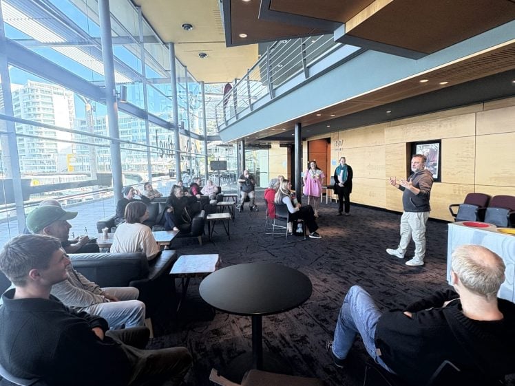 Image of A group of people sits and listens to a speaker standing near the front of a modern, glass-walled room with city views. Some attendees are seated at round tables; others stand near the entrance.