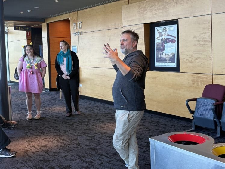 Image of A man in casual clothes speaks to a small group in a modern corridor with wooden panelled walls. Three people listen nearby. A recycling bin and a poster are visible in the background.