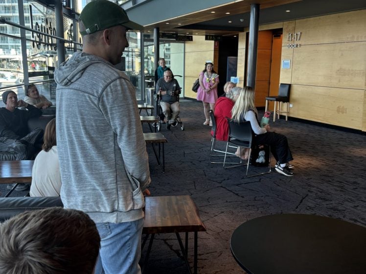 Image of People are sitting and standing in a modern waiting area with large windows, wooden walls, and upper-level railings. Some are talking whilst others appear to be waiting or relaxing.