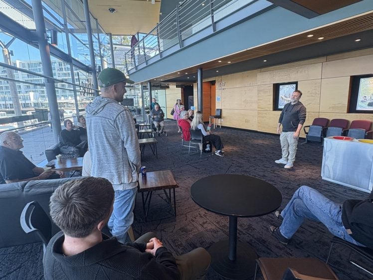 Image of People are seated and standing in a modern, spacious room with large windows, listening to a person speaking at the front near a screen. The atmosphere appears casual and conversational.