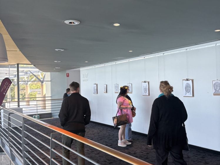 Image of Four people walk through a corridor art gallery with framed portraits hanging on the wall. The space is modern, with a metal railing and carpeted floor. Natural light streams in through large windows on the left.