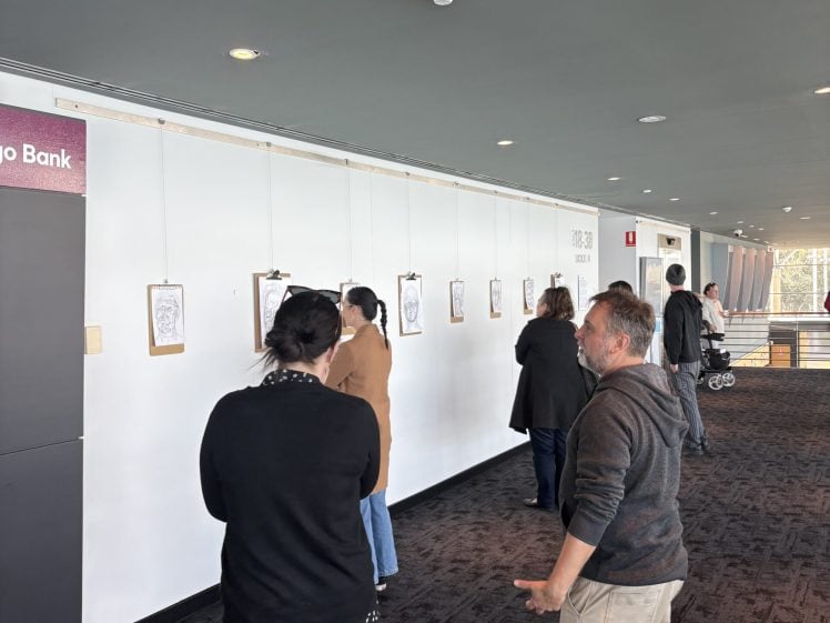 Image of A group of people observe framed artwork displayed on a white wall in a modern indoor gallery space with carpeted floors and large windows in the background.