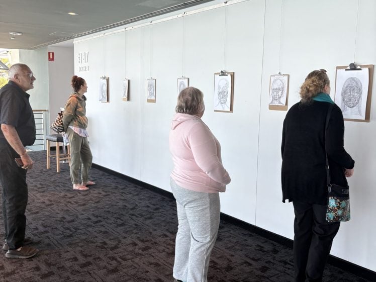 Image of Several people stand and walk along a white wall, viewing portrait drawings displayed in a gallery setting. The artworks are hung in a row at eye level, and the visitors appear to be observing the portraits closely.