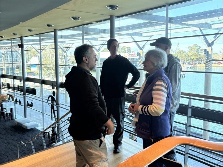 Image of Four people stand and talk on an upper floor of a modern building with large windows overlooking water and trees. Sunlight brightens the scene, and the mood appears casual and friendly.