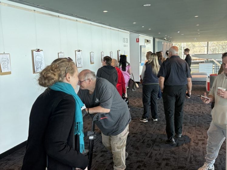 Image of People walk and look at framed artwork displayed on a white wall in a brightly lit corridor. Some people are talking, one uses a walking frame, and others look at the art or interact with each other.