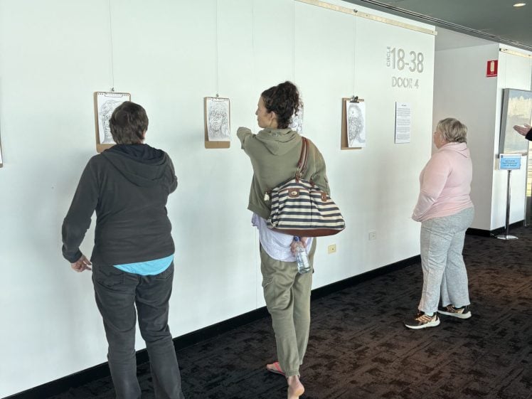 Image of Three people stand in front of a white wall, closely viewing drawings hung on clipboards in a gallery-style display. The setting appears to be an indoor public space with carpeted floors.