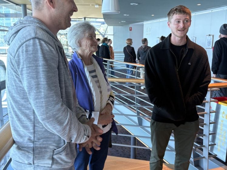 Image of Three people stand and chat on a staircase indoors. An older woman in a striped shirt and light jacket stands between two men; one in a grey hoodie and green cap, the other in a black jacket and green trousers. Sunlight streams in behind them.