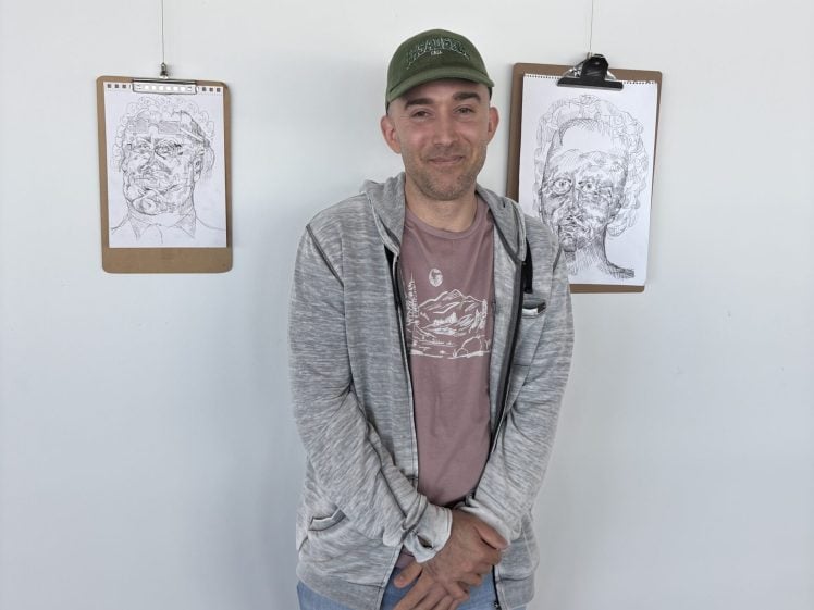 Image of A person wearing a green cap, grey hoodie, and jeans stands in front of a white wall with two clipboards holding black-and-white portrait sketches hanging behind them.