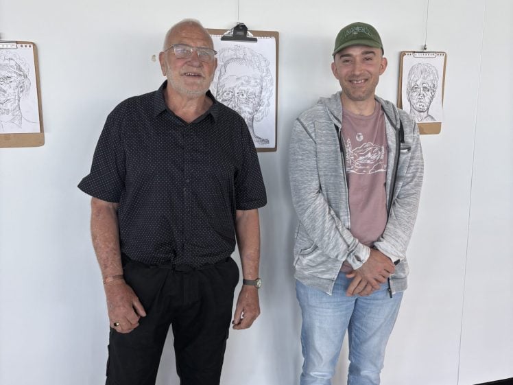 Image of Two men stand side by side in front of a white wall with clipboards displaying line drawings of faces behind them. One man is older with glasses, the other is younger wearing a cap and casual clothing.