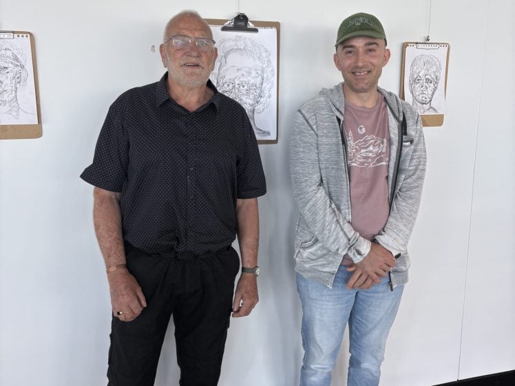 Image of Two men stand side by side indoors, smiling at the camera. Behind them, drawings of faces are displayed on clipboards attached to a white wall. The man on the left wears glasses and a black shirt; the man on the right wears a green cap and a grey jacket.