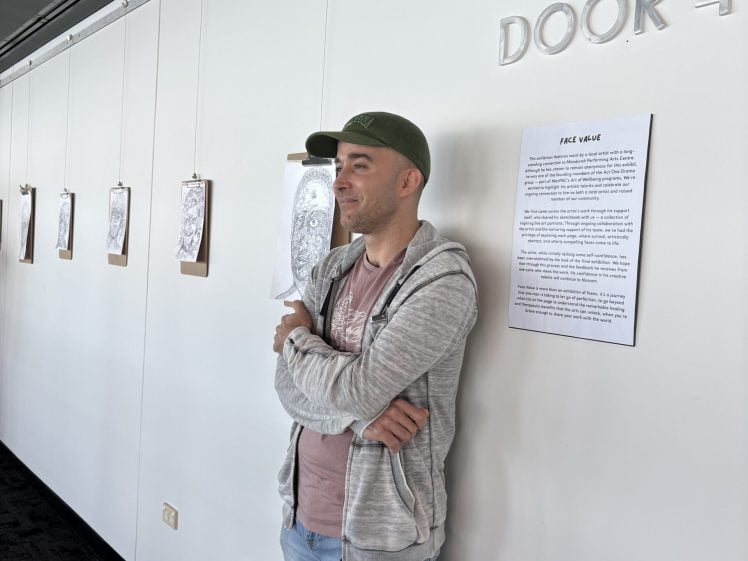 Image of A man in a green cap and grey hoodie stands with arms crossed, smiling in front of a white wall displaying several framed drawings and a printed description in a gallery-like setting.