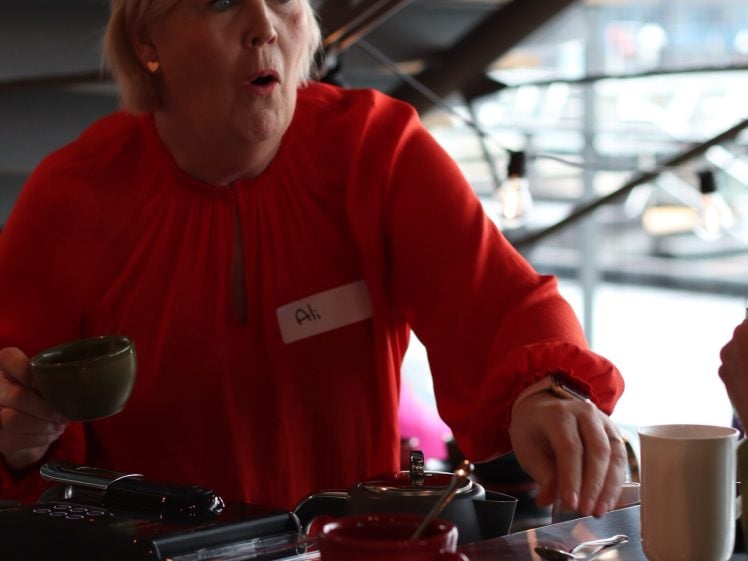 Image of A woman in a bright red blouse holding a green cup stands at a counter with coffee cups, appearing surprised or mid-conversation. She wears a name badge and is indoors with large windows in the background.
