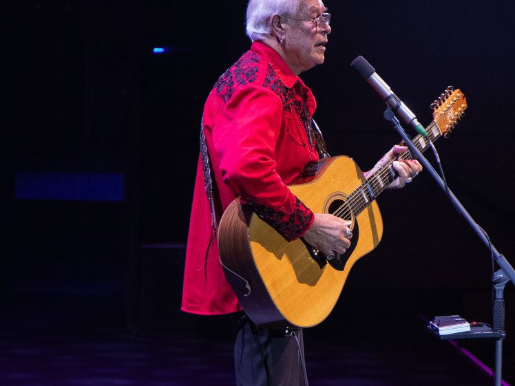 Image of An older man with white hair, wearing a red embroidered shirt, sings and plays an acoustic guitar on stage into a microphone, with a dark background.