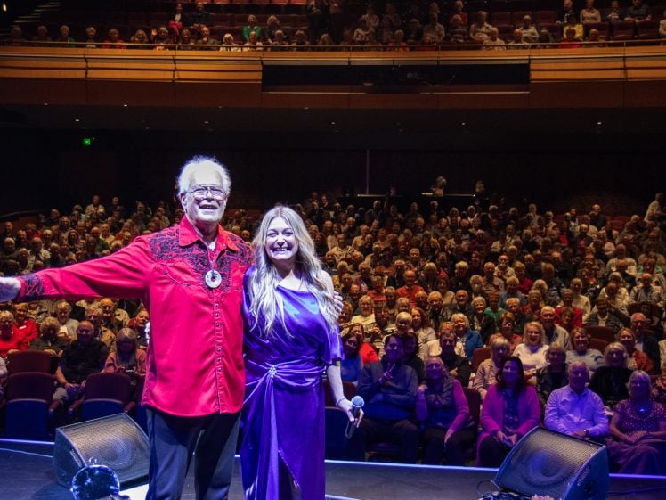 Image of Two performers, one in a red shirt and one in a purple dress, stand smiling on stage in front of a large seated audience in a theatre. The audience fills the background, watching the performers.