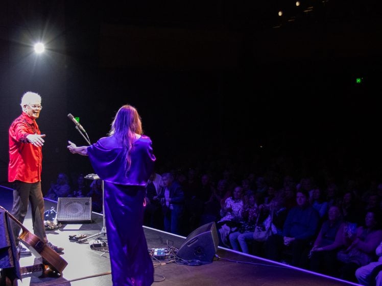 Image of Two performers stand on a stage facing a seated audience, one in a red shirt gesturing out, the other in a long dress with arms open. Stage lights shine brightly, illuminating the performers and part of the audience.