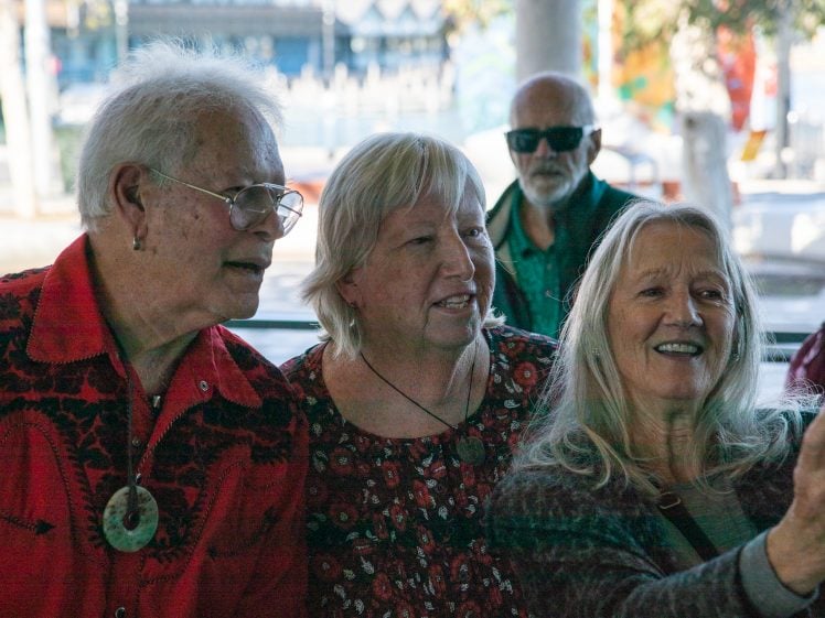 Image of Three older adults stand close together, smiling and taking a selfie outdoors. Another older man stands in the background, wearing sunglasses. The group appears cheerful and relaxed in a sunny setting.