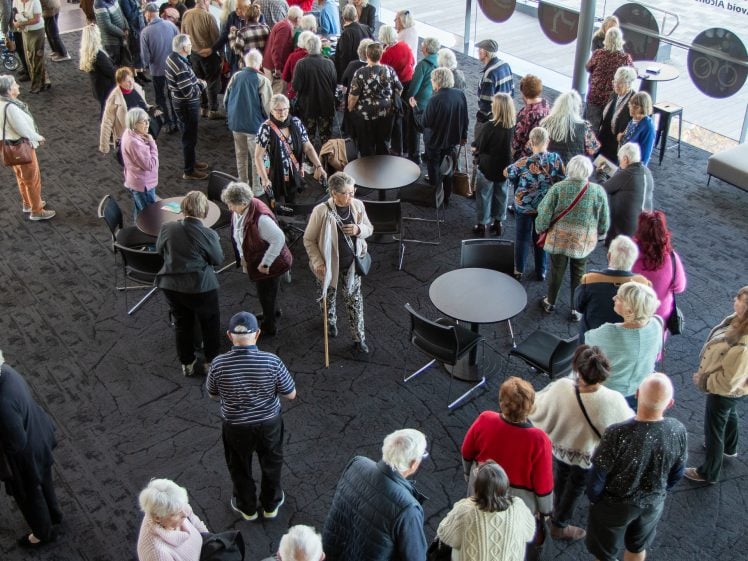 Image of A large group of elderly people, some with walking aids, gather in a spacious indoor area with tables and chairs, standing in queues and mingling near large glass windows.
