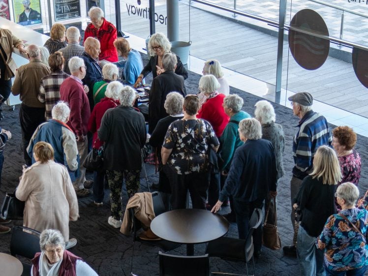 Image of A group of older adults, mostly with grey or white hair, gather indoors near tables and chairs, forming a queue or cluster by large windows with natural light streaming in.