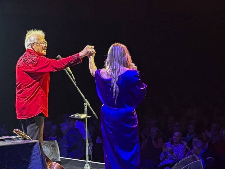 Image of An older man in a red shirt and a woman in a blue dress stand on stage facing an audience, holding hands and raising them together as if acknowledging applause. Musical instruments and microphones are visible nearby.