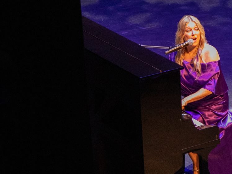 Image of A woman in a purple dress sings and plays piano on stage, illuminated by stage lights, with a microphone in front of her and a dark background behind the piano.