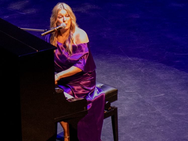 Image of A woman in a shiny purple dress sits at a black grand piano, singing into a microphone on a dimly lit stage with a blue and purple backdrop.