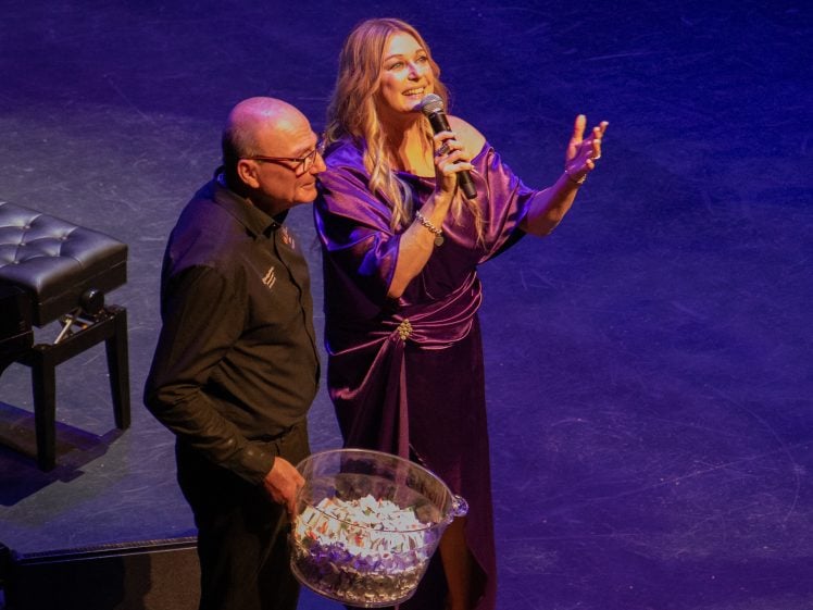 Image of A woman in a purple dress speaks into a microphone on stage, gesturing with her hand, while a man next to her holds a large transparent bowl filled with small pieces of paper.