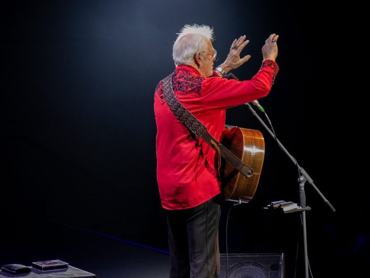 Image of An elderly man with white hair, wearing a bright red shirt and holding an acoustic guitar, stands on stage facing the audience with his arms raised, illuminated by stage lighting.