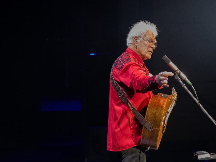 Image of An older man with white hair, wearing a red shirt with black embroidery, stands on stage playing an acoustic guitar and pointing whilst singing into a microphone under stage lights.