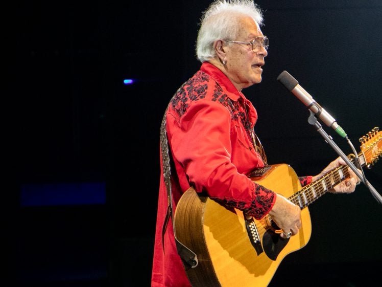 Image of An older man with white hair and glasses, wearing a red shirt with black patterns, plays an acoustic guitar and sings into a microphone on stage.