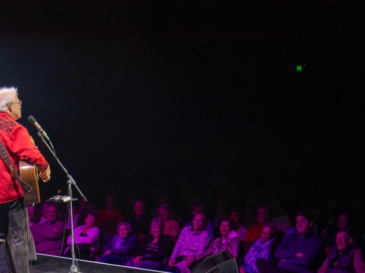 Image of An older musician in a red shirt plays guitar and sings on stage, facing a smiling and laughing audience seated in a dimly lit theatre.