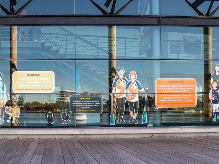 Image of Illustrated figures with diverse abilities and backgrounds are displayed on large glass windows, accompanied by colourful panels of text describing their stories. The waterfront and promenade are visible outside the building.