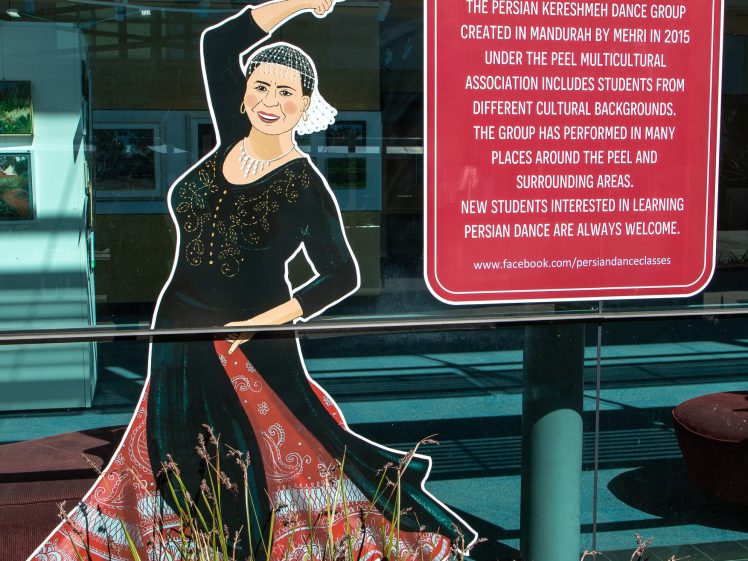 Image of A colourful illustration of a woman in traditional Persian dance attire is displayed next to a sign describing the Persian Kereshmeh Dance Group, founded by Mehri Kalani in Mandurah in 2015. Grass grows in front of the display.