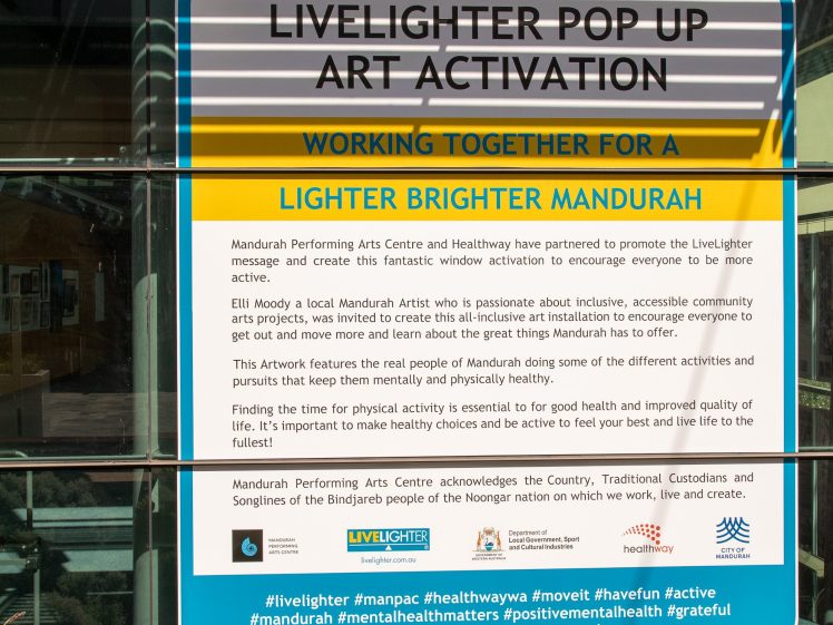 Image of A large informational sign on a glass window promotes the Livelighter Pop Up Art Activation, describing its goal to support community mental health and wellbeing in Mandurah. Several hashtags appear at the bottom.