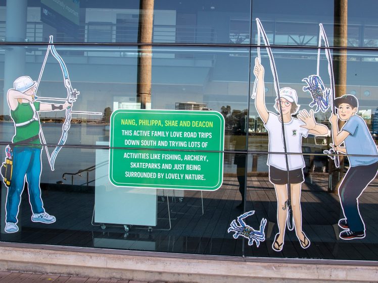 Image of Illustrations of four people holding bows and arrows are displayed on a glass wall next to a sign describing their love of fishing, archery, skateparks, and road trips, set against a reflective outdoor backdrop.