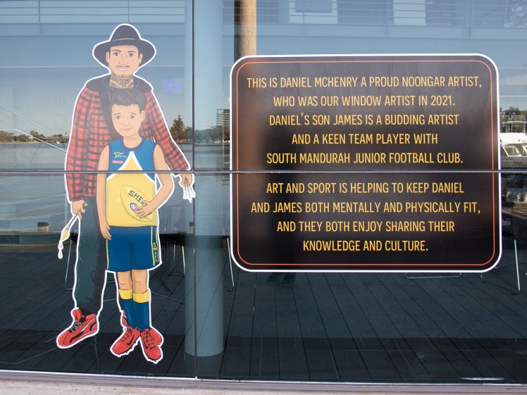 Image of A cardboard cut-out of two boys stands next to a large sign describing Daniel McHenry, a Noongar artist, and his son James, a young footballer, highlighting their connection to art, sport, and cultural sharing.