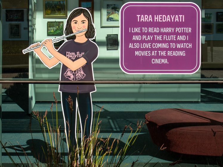 Image of A cardboard cut-out of a girl playing the flute stands in front of a window with art displayed inside. A sign reads: Tara Hedayati: I like to read Harry Potter and play the flute and I also love coming to watch films at the Reading Cinema.