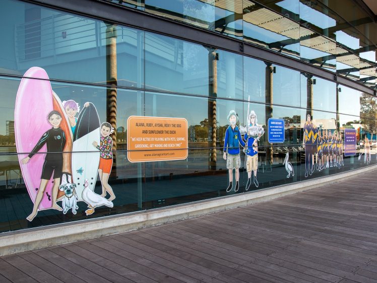 Image of Colourful illustrations of children in various uniforms and sports outfits are displayed on a large glass window along a wooden walkway, with water and buildings visible in the reflection.