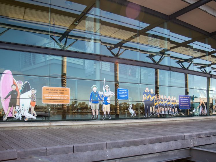 Image of A large glass window features colourful illustrations of surf lifesavers, lifeguards, a dog, and signs, with wooden decking and reflections of the waterfront in the background.