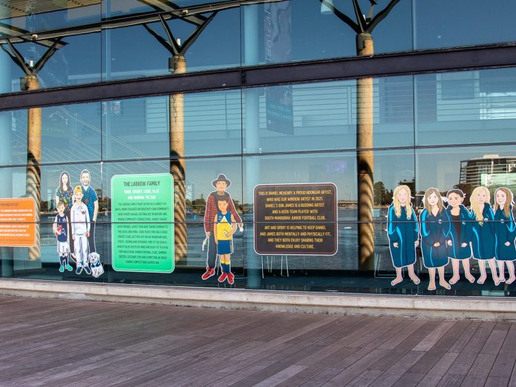 Image of A glass building façade displays colourful illustrated cut-outs of diverse people in various uniforms and attire, with informational panels beside them. Reflections of nearby structures and trees are visible in the glass.