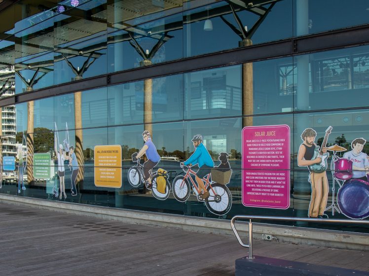 Image of Illustrated figures of people cycling, playing musical instruments, and engaging in activities are displayed on large glass windows of a modern building along a wooden walkway by the water.